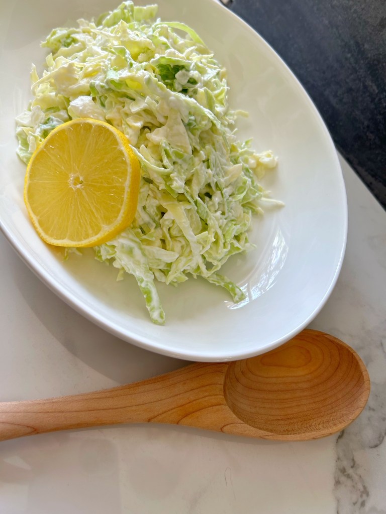 This is an image of cabbage salad.  The cabbage salad is in a bowl with a round slice of lemon and there is a wooden spoon beside the bowl.