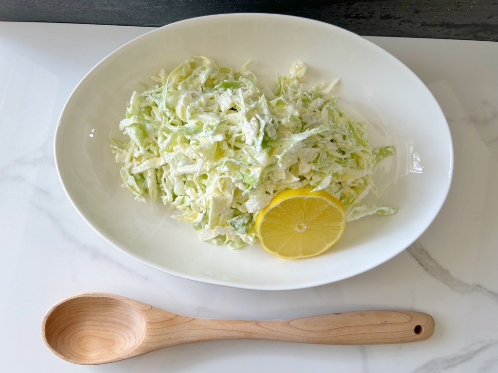 This is an image of cabbage salad.  The cabbage salad is in a bowl with a round slice of lemon and there is a wooden spoon beside the bowl.
